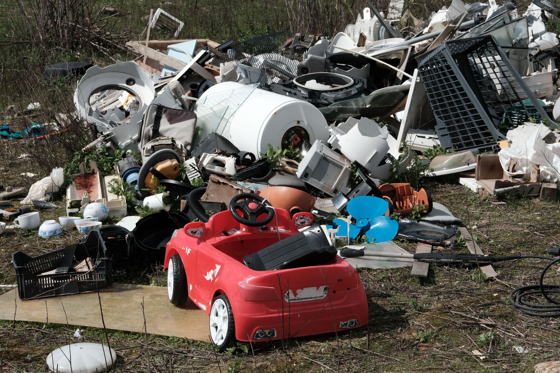 A red toy car sits among discarded appliances and debris, symbolizing environmental neglect.